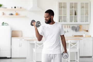 Smiling millennial handsome bearded african american male athlete in white clothes lifts dumbbells in minimalist kitchen