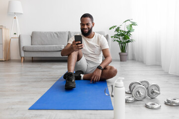 Smiling millennial african american bearded sit on mat on floor in room interior with dumbbells, bottle of water