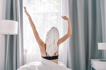 Fototapeta na wymiar Happy woman in black top and towel sits in bed waking up from adequate and healthy sleep, feeling good stretching arm muscles after sleep and long immobility wakes up to start new day with smile