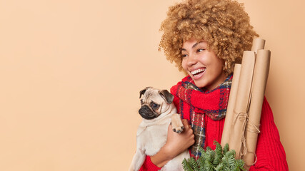 Horizontal shot of positive carefree curly woman poses with pedigree pug dog holds items for decoration gets ready for winter holidays looks away poses against beige background with copy space