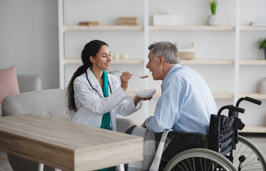 Obraz premium Medical assistance for senior patients with disability. Young nurse feeding to older Caucasian man in wheelchair at home