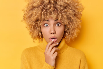 Portrait of stunned young curly woman gasps from astonishment feels stupefied keeps hand on chin opens mouth dressed in casual jumper isolated over yellow background. Human reactions concept