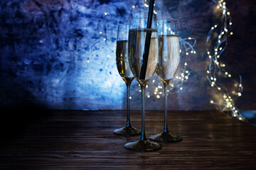 New year celebration with sparkeling champagne. Champagne glasses with drinking straw on wooden bar counter against luminous blue background with fairy lights.