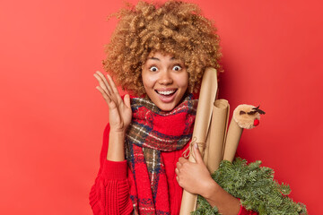 Optimistic woman with curly hair raises palm feels excited holds spruce wreath and rolled paper feels glad to celebrate Christmas holidays isolated over red background. Winter time and celebration