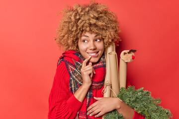 Dreamy cheerful young woman anticipates winter holidays holds festive Christmas wreath and rolled paper looks away dressed in warm knitted sweater scarf around neck isolated over red background