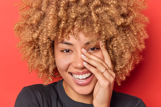 Close up portrait of happy curly haired woman keeps hand on face smiles broadly expresses sincere feelings looks with eyes full of joy isolated over red background. Positive emotions concept