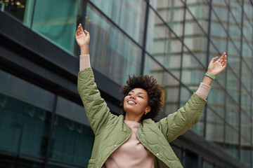 Carefree joyful woman dressed in casual jacket raises arms poses against modern glass buildings in...