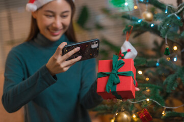 Happy Asian woman using smartphone mobile taking photos of Christmas gift box with Christmas tree in background