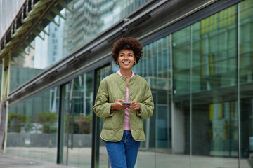 Young modern woman with curly hair dressed casually uses smartphone for messaging during walking in city uses modern technology device poses against modern business centre. Lifestyle concept - Powered by Adobe
