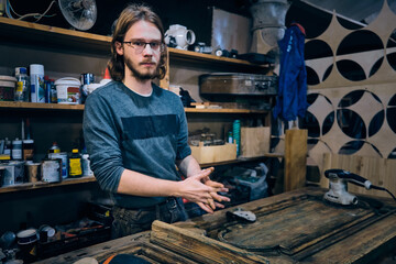 Portrait of young adult man at carpenter workshop. Man works on vintage wooden door restoration at carpenter workshop. Small business. Handicraft enterprise. Moody low key photo