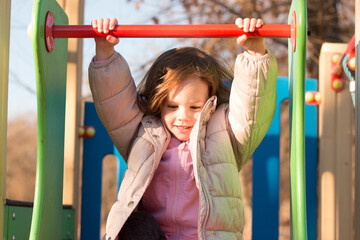 Fototapeta premium Small child with long red hair against the background of a blue sky in warm clothes on the playground