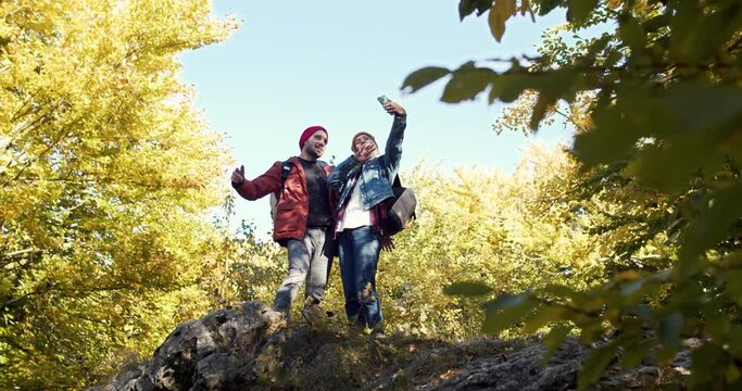 Joyful couple of man and woman standing in forest and making selfie photos on cellphone. Beautiful female and happy male posing while taking pictures on smartphone. Hiking and tourism concept