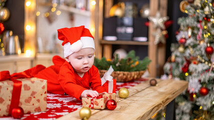 baby girl in red costume lying on wooden surface with Christmas decorations. baby lies on its tummy, next to gift boxes during Christmas celebration. Carefully studying world around you. Christmas eve