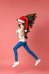 happy teenage girl in knitted sweater, full-length, wearing a Santa hat on her head, with Christmas tree decorated with red Christmas balls on her shoulder, walks on pink background. Dynamic image.