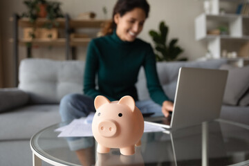 Close up of piggy bank on table, happy Hispanic woman manage household budget pay online on laptop...