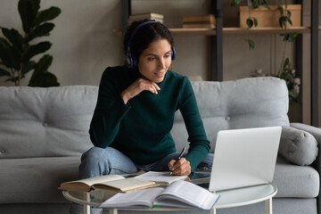 Millennial Latino woman in headphones make note write studying online on laptop at home. Young Hispanic female student busy working on internet on computer, take distant course. Education concept.