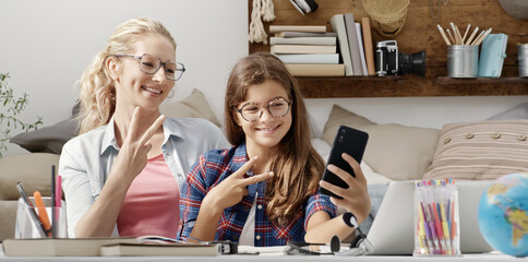 Mother and her young teenager student daughter smiling together use smartphone and show peace v sign with hand gesture, sitting at desk in home, happy family life and modern communication concept