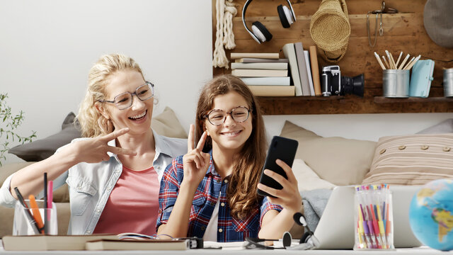Mother and her young teenager student daughter smiling together use smartphone and show peace v sign with hand gesture, sitting at desk in home, happy family life and modern communication concept
