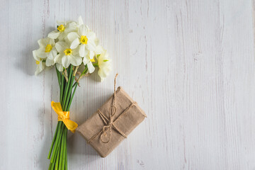 Spring holiday greeting card; bouquet of white daffodils and handmade gift box on white wooden background; copy space