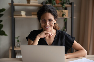 Smiling young Latino woman in glasses look at laptop screen work online on gadget at home office....