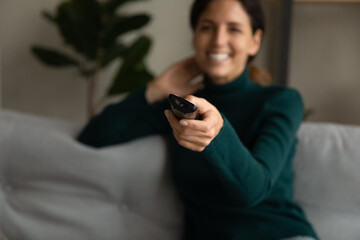 Fototapeta premium Crop close up of smiling Hispanic woman relax at home watch TV switch channel with remote control. Happy Latin female rest have fun enjoy television program online. Technology, entertainment concept.
