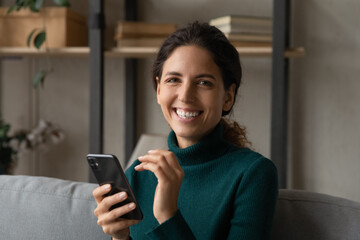 Portrait of happy millennial Latino woman relax on couch at home use modern cellphone gadget texting messaging online. Smiling young Hispanic female client or user browse web on smartphone device.