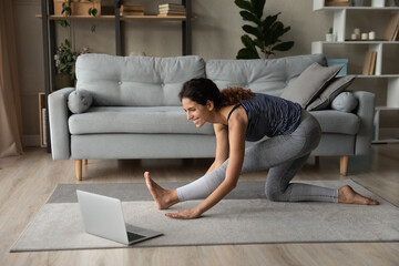 Happy millennial sporty woman do morning exercise or gymnastics watching video online class on laptop. Smiling young Latino female do pilates train with coach on webcam call on computer from home.
