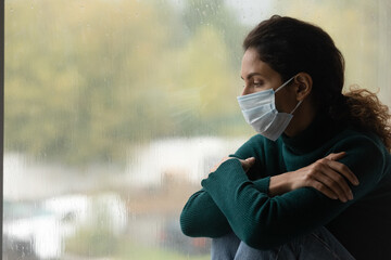 Unhappy sick young Latino woman wear facemask look in window distance feel lonely on lockdown....