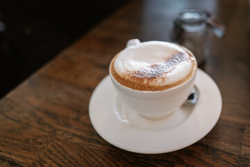 Shallow focus on the froth of a cappuccino coffee in a white cup and saucer. A teaspoon is in the saucer and it is placed on a wooden table. A glass sugar bowl is behind.