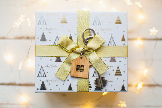 House key with keychain cottage. Happy New Year-golld bow in gift box, background with snow, fairy lights, greeting card. Purchase, construction, relocation, mortgage. Merry Christmas