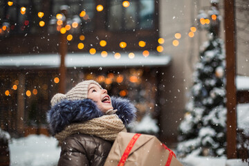 Happy girl with a gift box for Christmas catches snowflakes with his tongue on a city in winter with snow on festive market with decorations and lights. Warm clothes, knitted hat, scarf and fur. 