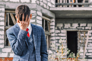 Obraz premium distressed young man in fashionable suit stands on the background of destroyed unfinished house, insolvent person to pay for housing or mortgage, construction or mortgage crisis concept 