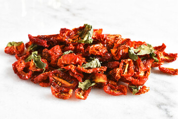 A close up image of sun dried cherry tomatoes with dried organic basil leaves on a white marble table top. 