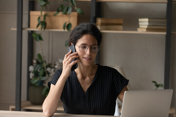Young Hispanic woman sit at desk at home office work online on laptop talk on smartphone with client or customer. Confident Latin female use computer have cellphone communication. Technology concept.