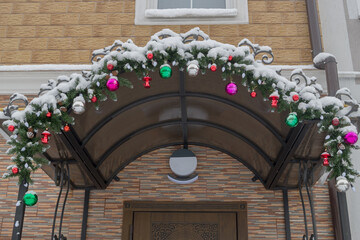Brown door to entrance to residential apartment building, with decorative yellow facade. Metal canopy decorated with fir branches, garlands and Christmas tree toys dusted with snow is installed above.