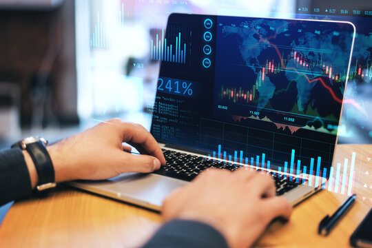 Close up of businessman hands at wooden desktop using laptop with glowing forex chart trading interface on blurry background. Market, economy and data exchange concept. Double exposure.