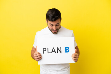 Young handsome caucasian man isolated on yellow background holding a placard with the message PLAN B