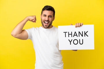 Young handsome caucasian man isolated on yellow background holding a placard with text THANK YOU and doing strong gesture