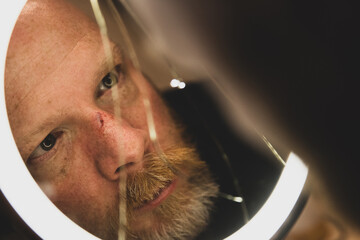 Middle aged man with a beard stares into a cracked mirror looking at the reflection the broken and bloody nose on his face. 