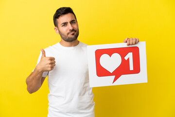 Young handsome caucasian man isolated on yellow background holding a placard with Like icon with thumb up