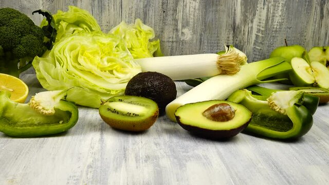 Vegetables and fruits of green color whole and halves lie on a wooden table.  Avocado and lemon, leek, broccoli, kiwi, apples and green bell pepper iceberg lettuce are on a table.