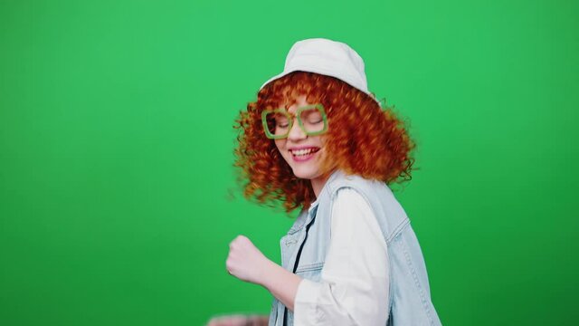 Young positive curly redhead lady in eyeglasses and panama hat walking and dancing, gesturing gotcha to camera