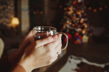 Woman relaxing with a cup of spiced tea in front of a fireplace in winter.Hands in a warm sweater are holding a mug of tea, close-up. Trying to warm my hands with a hot drink.