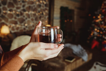 Woman relaxing with a cup of spiced tea in front of a fireplace in winter.Hands in a warm sweater are holding a mug of tea, close-up. Trying to warm my hands with a hot drink.