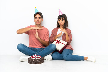 Young mixed race couple celebrating a birthday sitting on the floor isolated on white background making stop gesture with her hand to stop an act