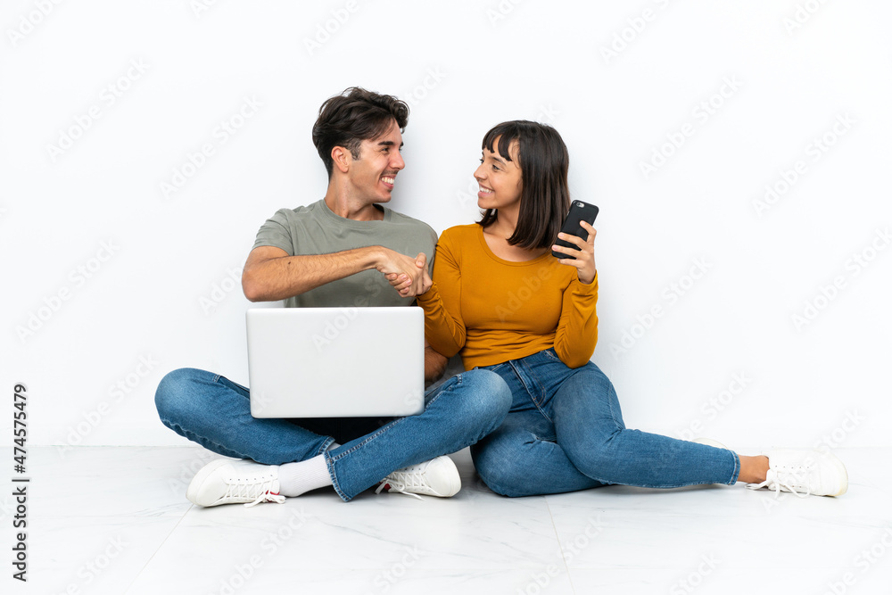 Wall mural young couple with a laptop and mobile sitting on the floor handshaking after good deal