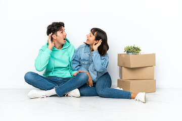 Young couple making a move while picking up a box full of things sitting on the floor isolated on white background listening to something by putting hand on the ear