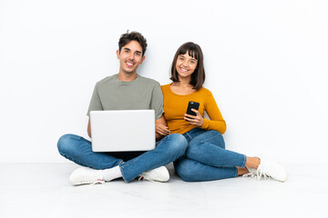 Young couple with a laptop and mobile sitting on the floor posing with arms at hip and smiling