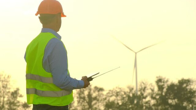 Windmill produces energy against cloudy sky. Focus changes from rotating turbine to engineer with tablet and portable radio looking at built station