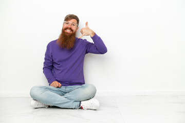Young caucasian reddish man sitting on the floor isolated on white background making phone gesture. Call me back sign